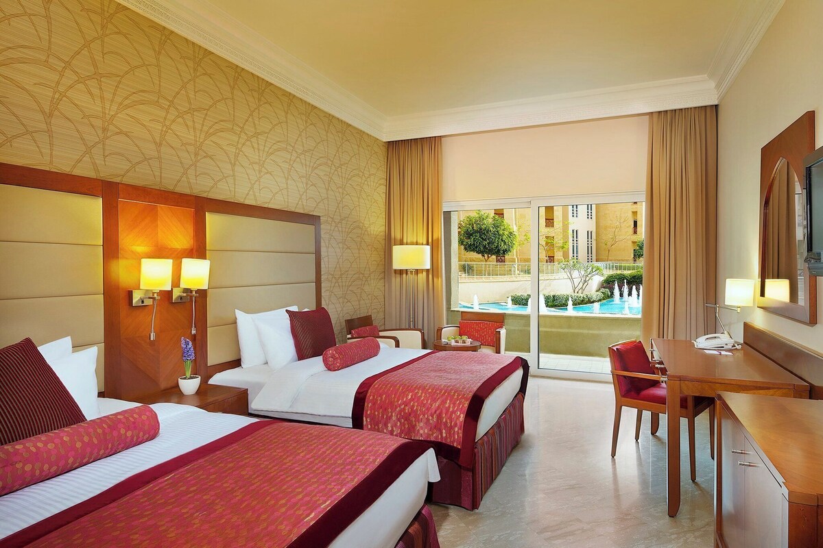 Habitación de hotel con dos camas, un escritorio, una silla, un televisor y vista a una piscina y edificios.