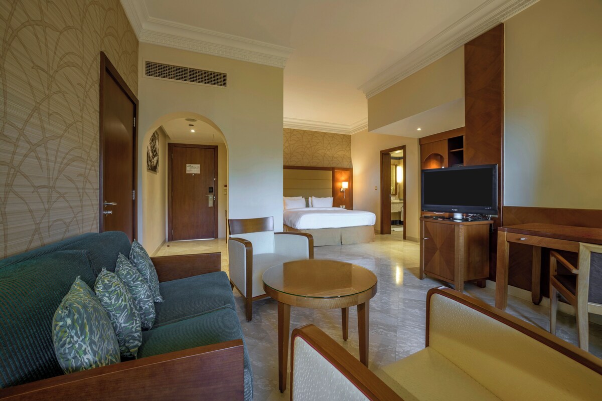Habitación de hotel con sofá, sillón, una pequeña mesa redonda, televisor, cama y escritorio.