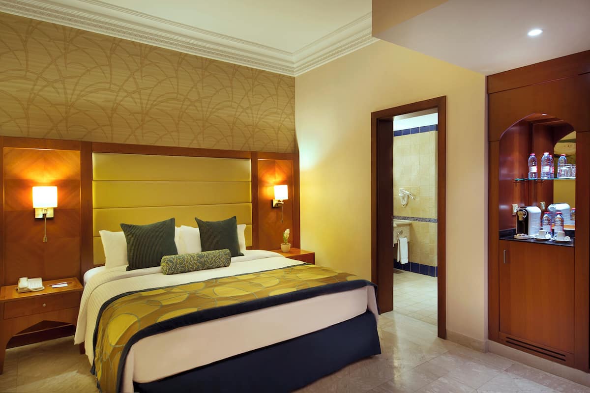 Habitación de hotel con una cama grande, dos mesitas de noche con lámparas, un armario de madera y un baño visible a través de una puerta abierta.