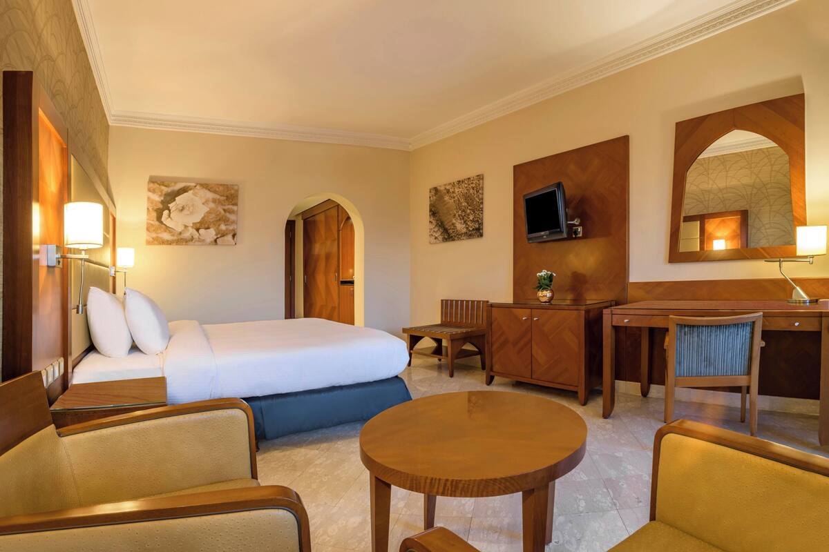 A hotel room with a bed, two armchairs, a wooden desk, a round table, and a television.