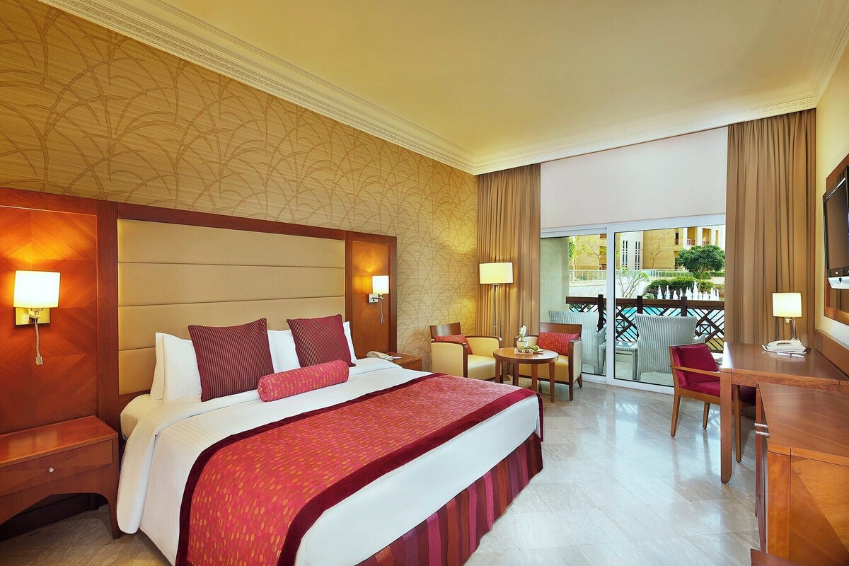Habitación de hotel con una cama grande, un escritorio, una silla, una mesita y un balcón con vista a la piscina.