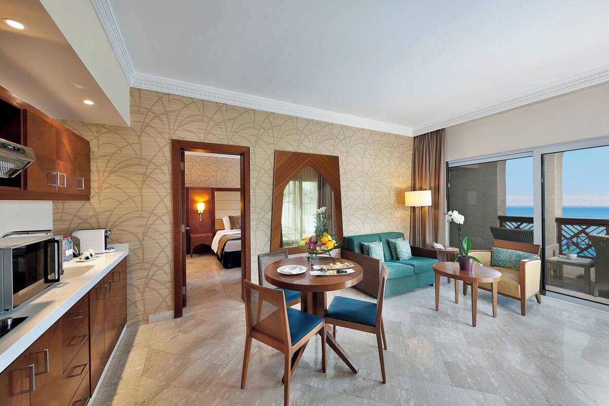 A hotel room with a dining area, a kitchen, a living area with a sofa and chairs, and a balcony with a view.