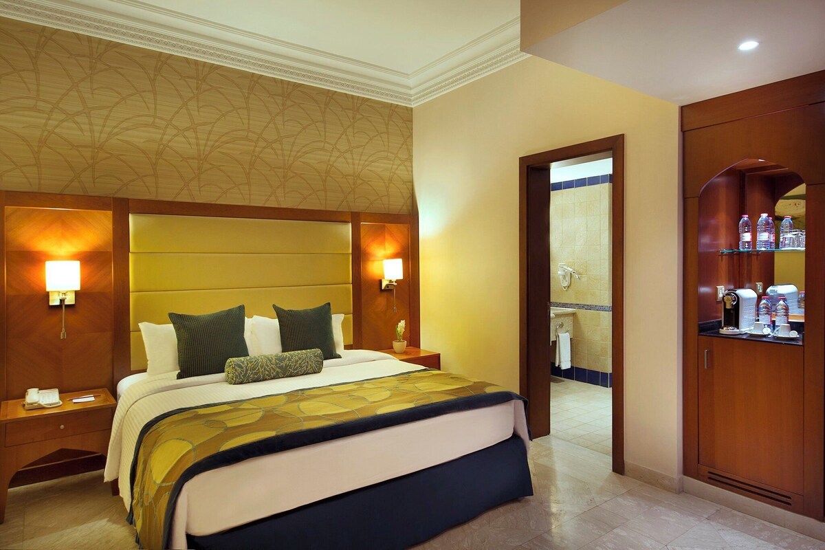 Habitación de hotel con una cama grande, lámparas de noche, mesita de noche, baño visible a través de una puerta abierta y un minibar.