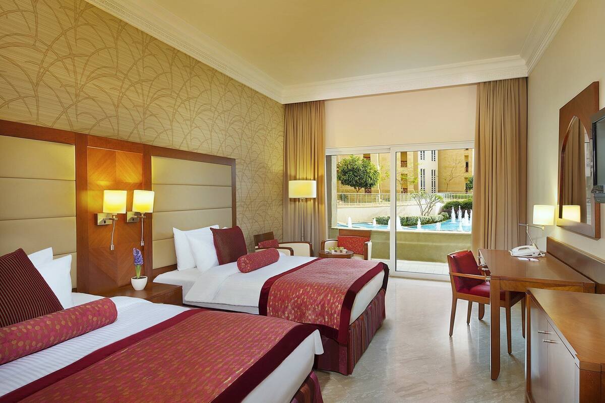 A hotel room with two beds, a desk, a chair, a TV, and a view of a pool and buildings.