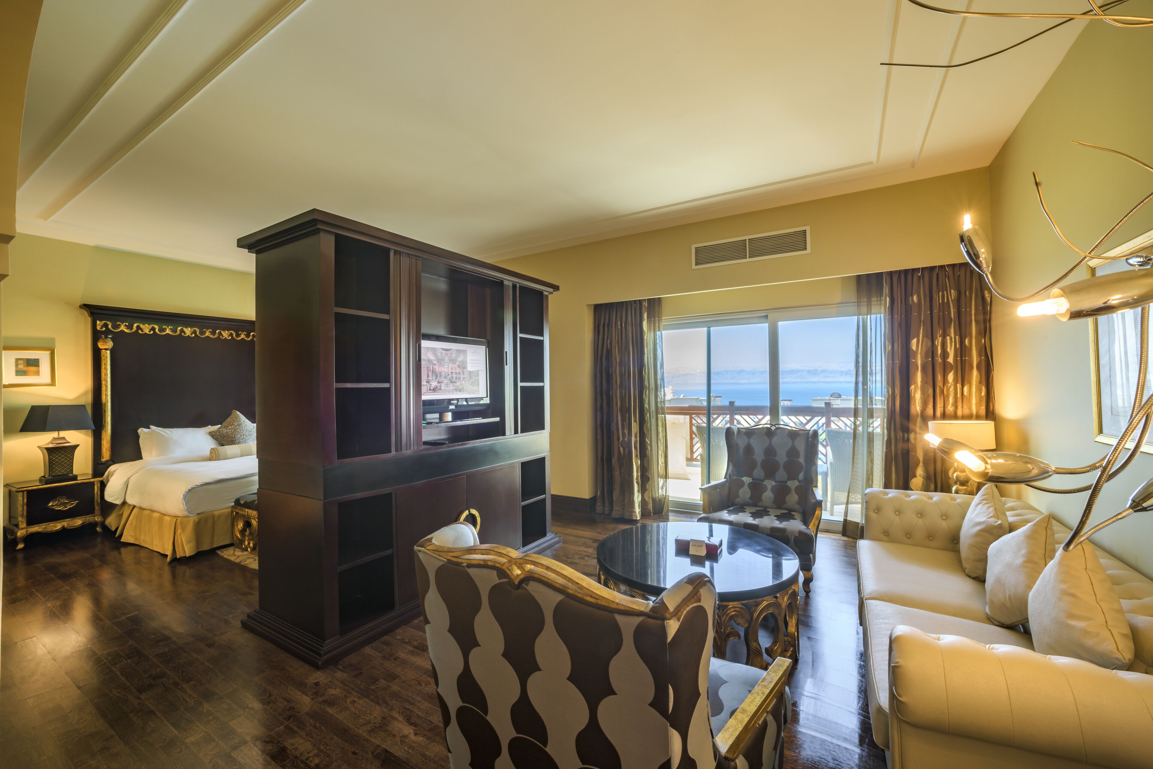 A hotel room with a bed, a large dark wooden cabinet, a round table, a sofa, and a view of the sea through large windows.