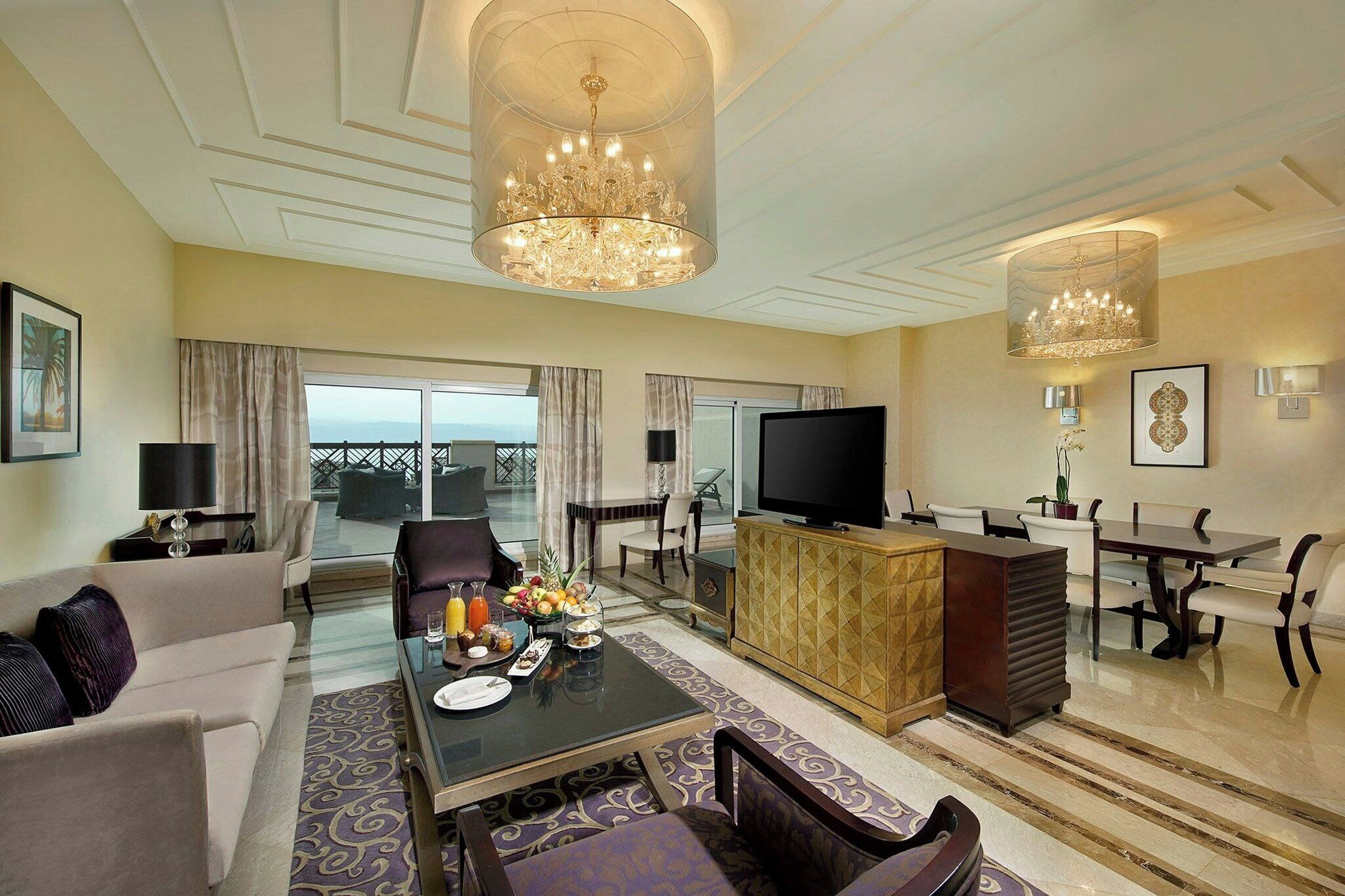 A spacious hotel living room with a large chandelier, a flat-screen TV, a dining area, and a balcony with a view.