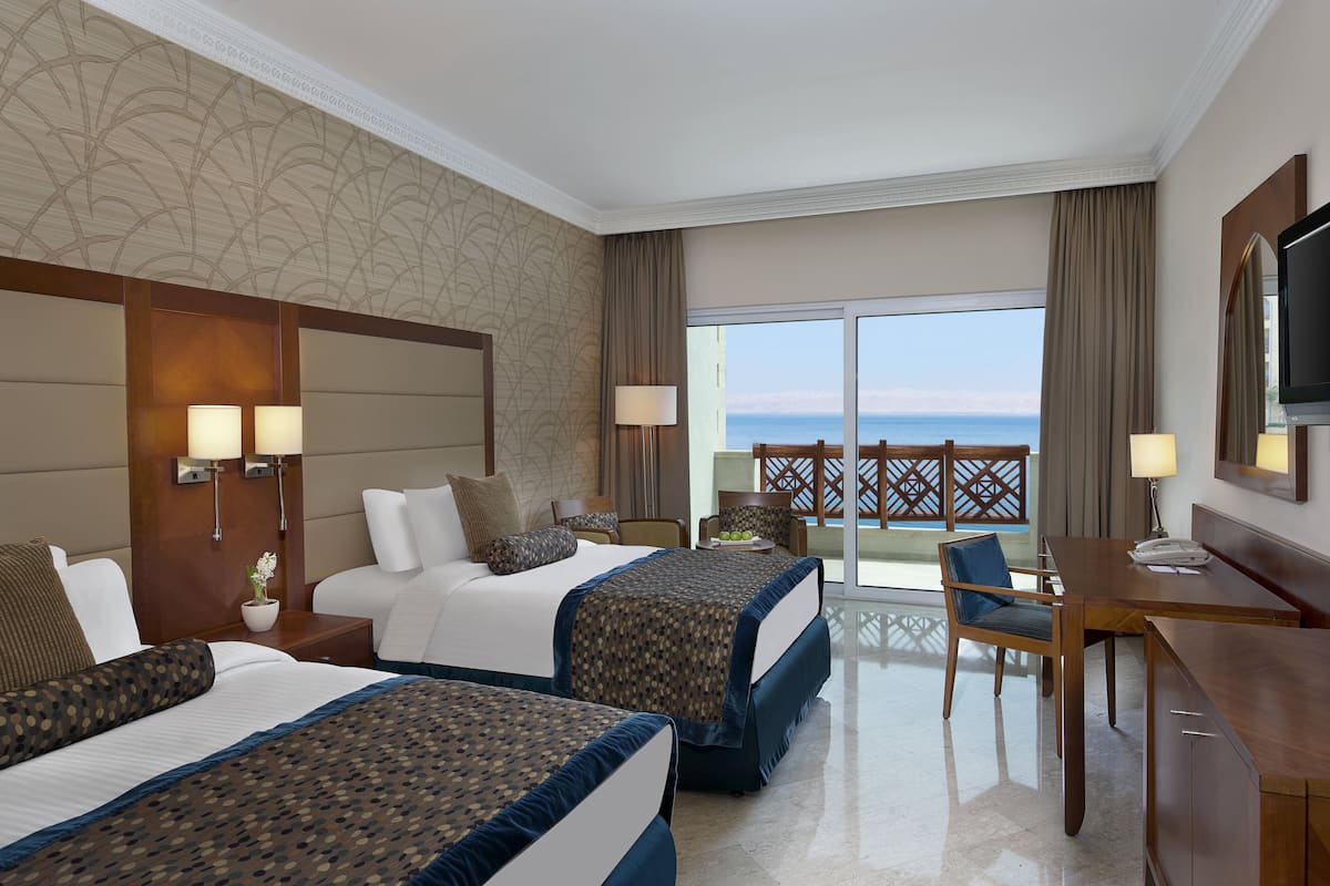 A hotel room with two beds, a desk, a chair, a TV, and a balcony with a view of the sea.