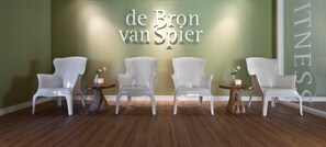 A reception area with four white chairs and small round tables, each with a candle. The wall behind has a sign that reads "de Bron van Spier". - Van Der Valk Hotel Spier - Dwingeloo (Spier)