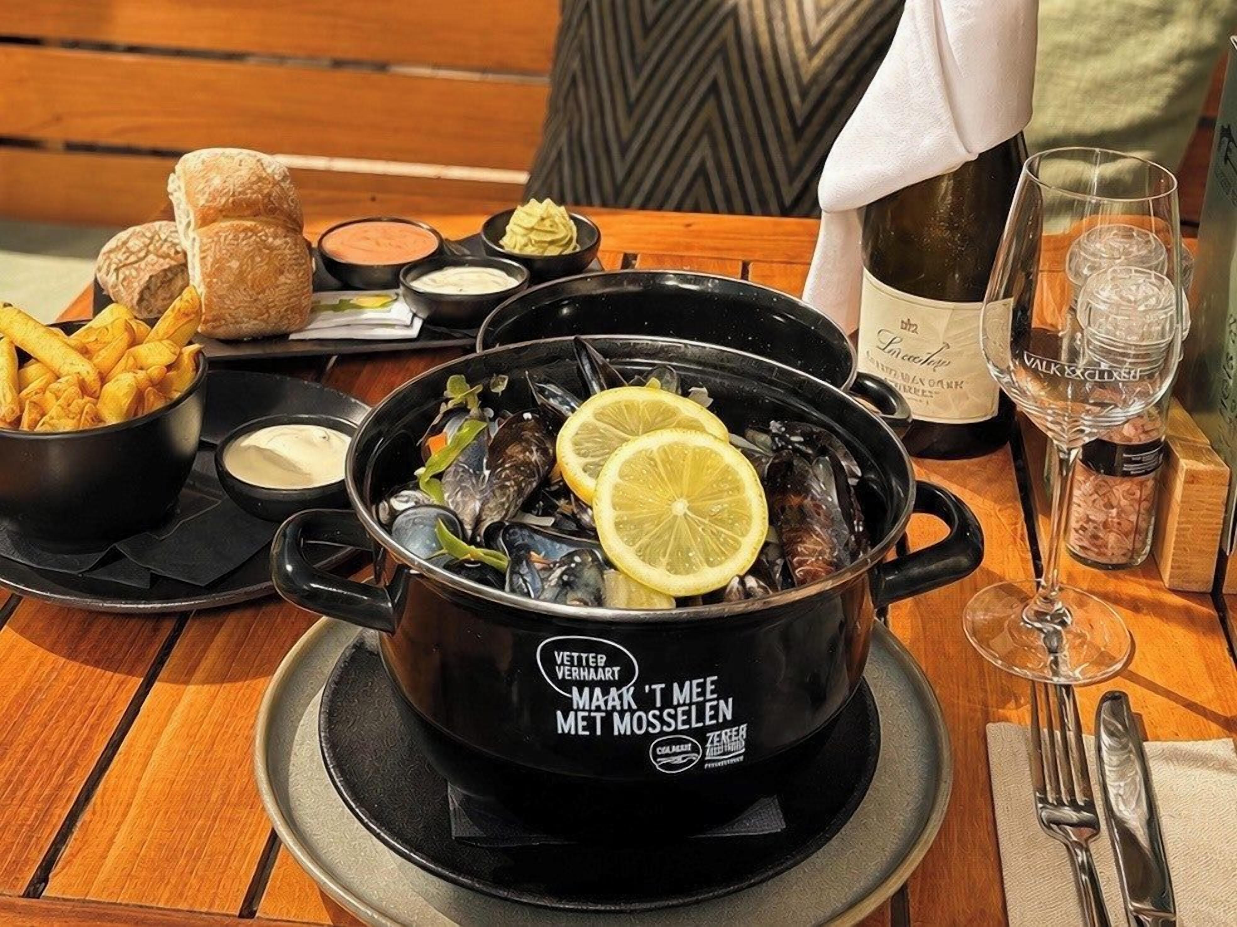 A dining table with a black pot of mussels, bread rolls, and a bottle of wine.