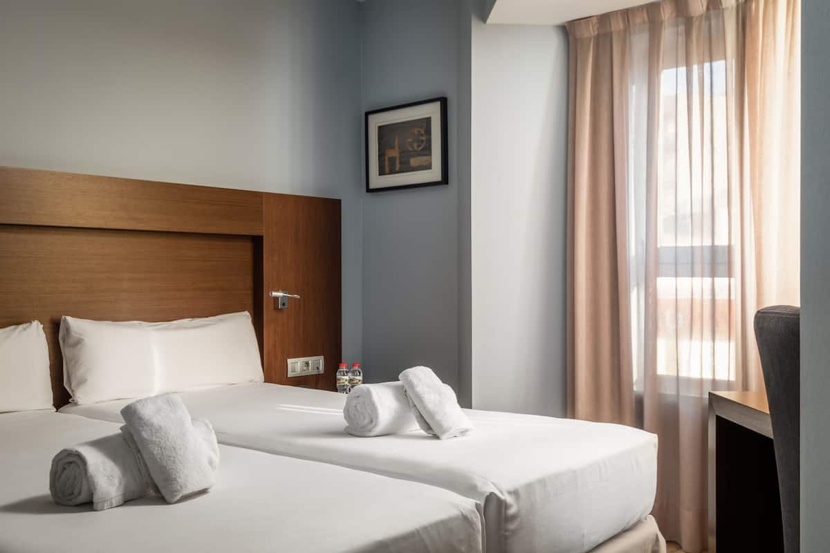 Une chambre d’hôtel avec deux lits, une tête de lit en bois, un tableau encadré au mur et une fenêtre avec des rideaux.