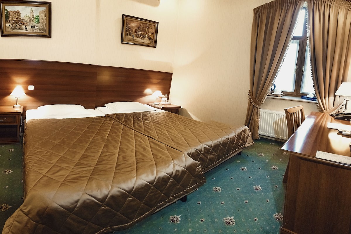 A hotel room with a large bed, two bedside tables with lamps, a wooden nightstand, a chair, a desk with a computer, and a window with curtains.