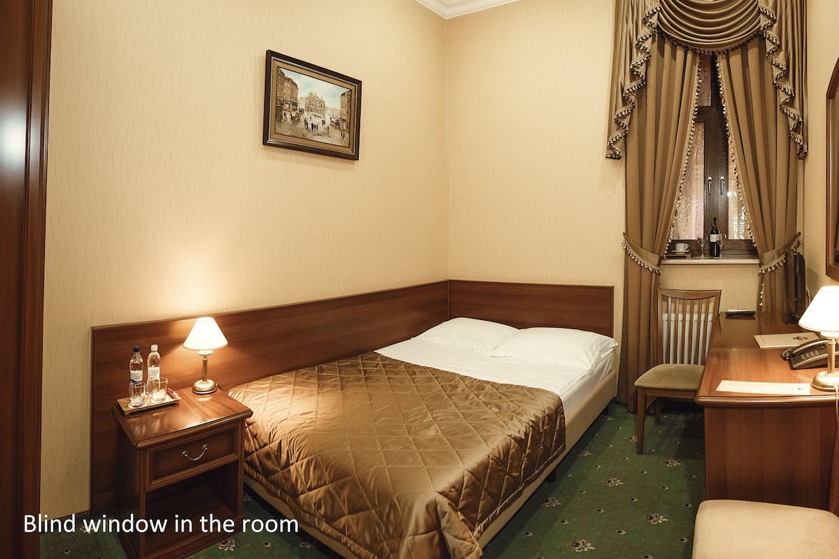 A hotel room with a bed, bedside table, chair, desk, lamp, and a window with curtains.