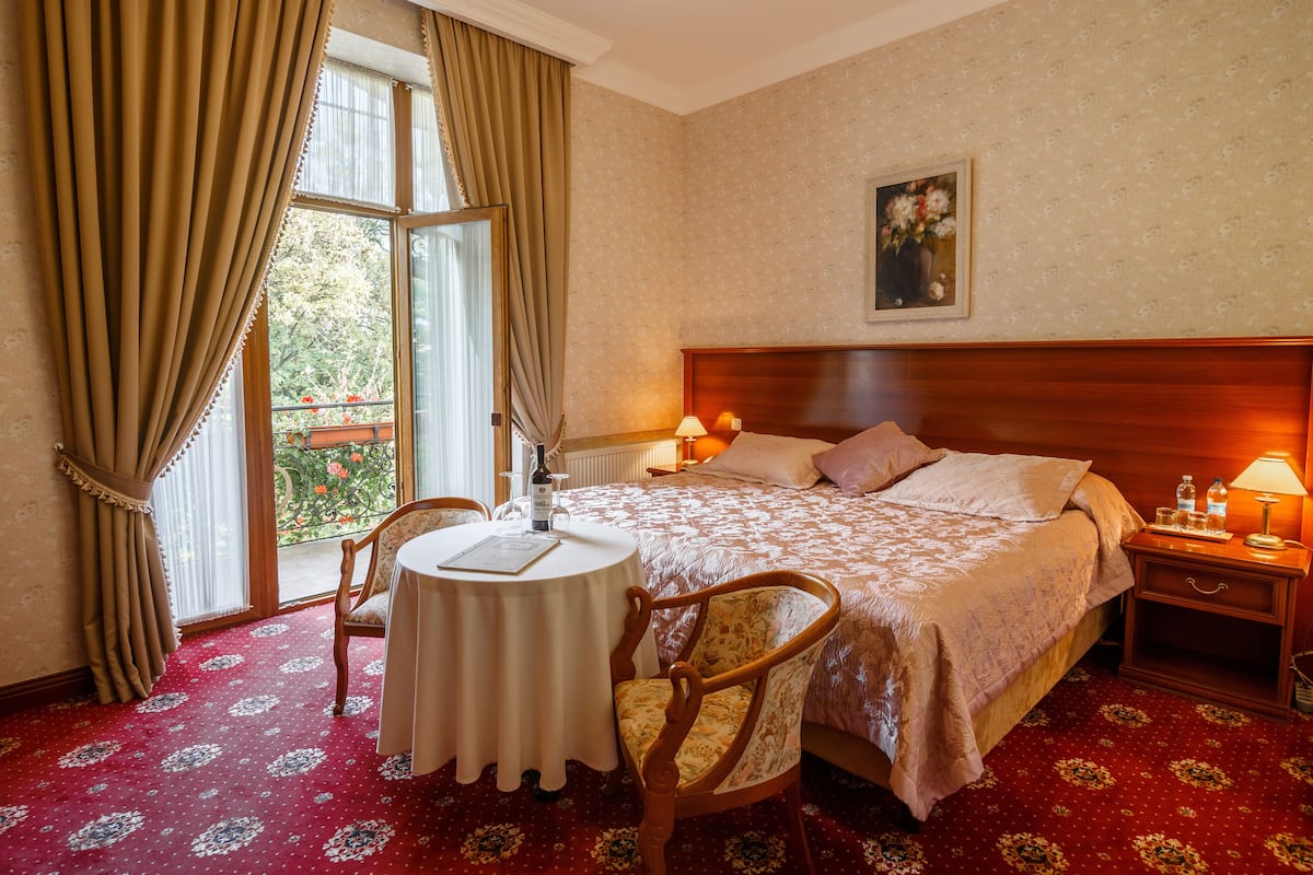 A hotel room with a large bed, a round table with a chair, a nightstand with a lamp, and a window with curtains.