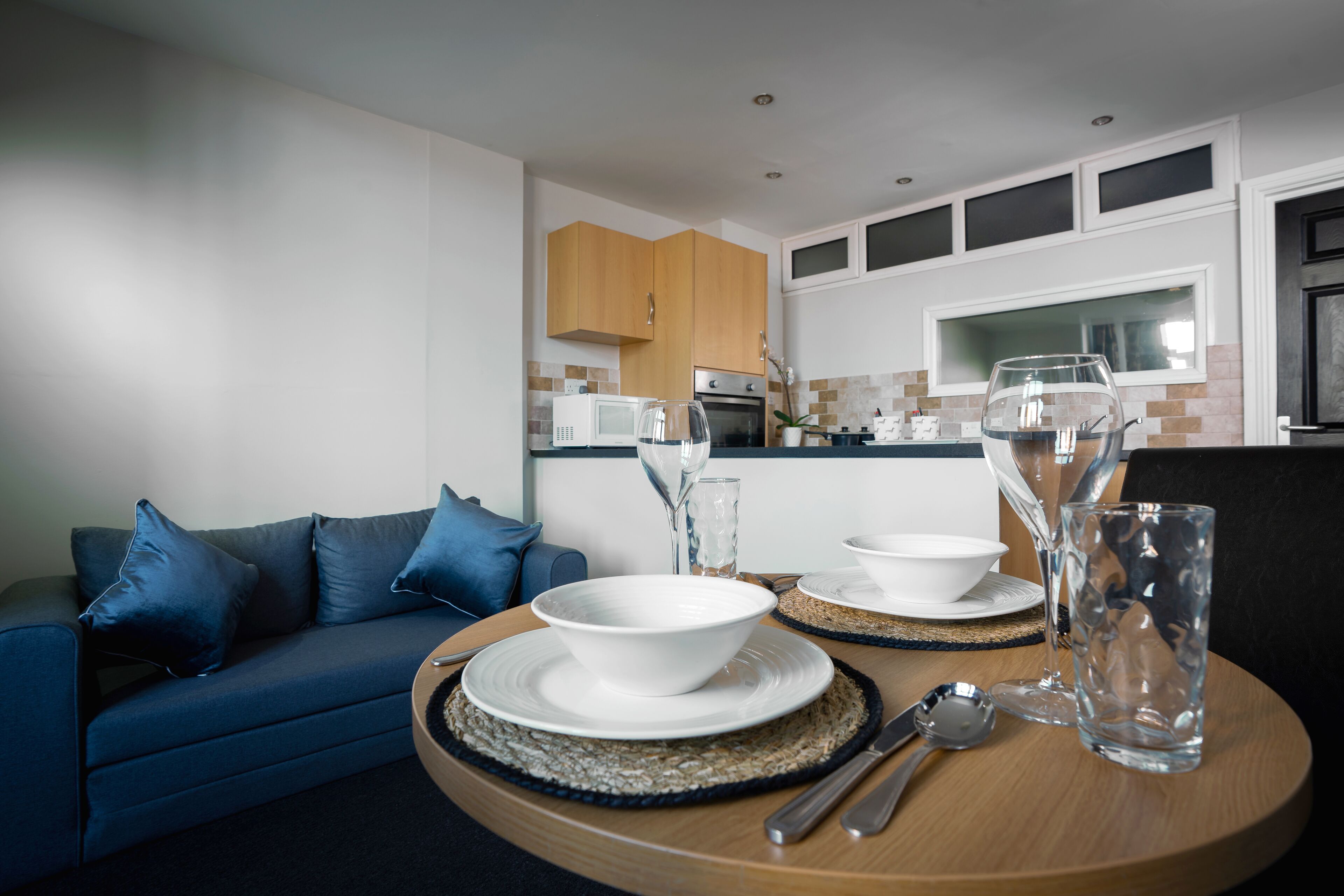 A dining area with a round table set for two, a blue sofa, and a kitchen in the background.