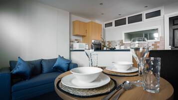 A dining area with a round table set for two, a blue sofa, and a kitchen in the background.
