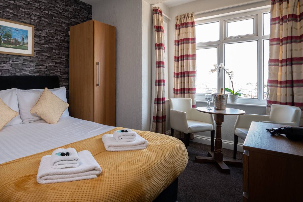 A hotel room with a stone wall, a bed with white linens, a wooden wardrobe, a dining table with chairs, and a window with curtains.