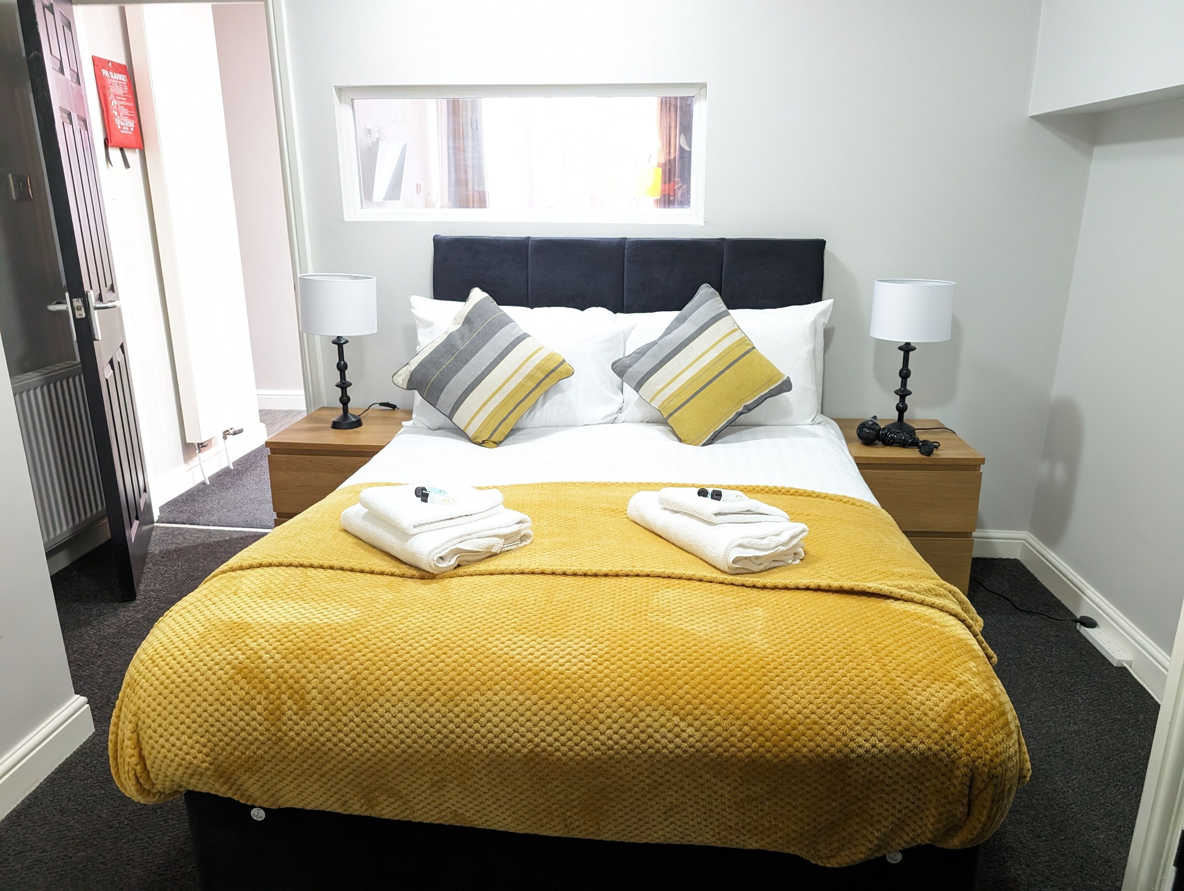 A neatly made bed with a yellow blanket, two pillows, and two folded towels. There are two bedside tables with lamps, a window with curtains, and a door with a red sign.