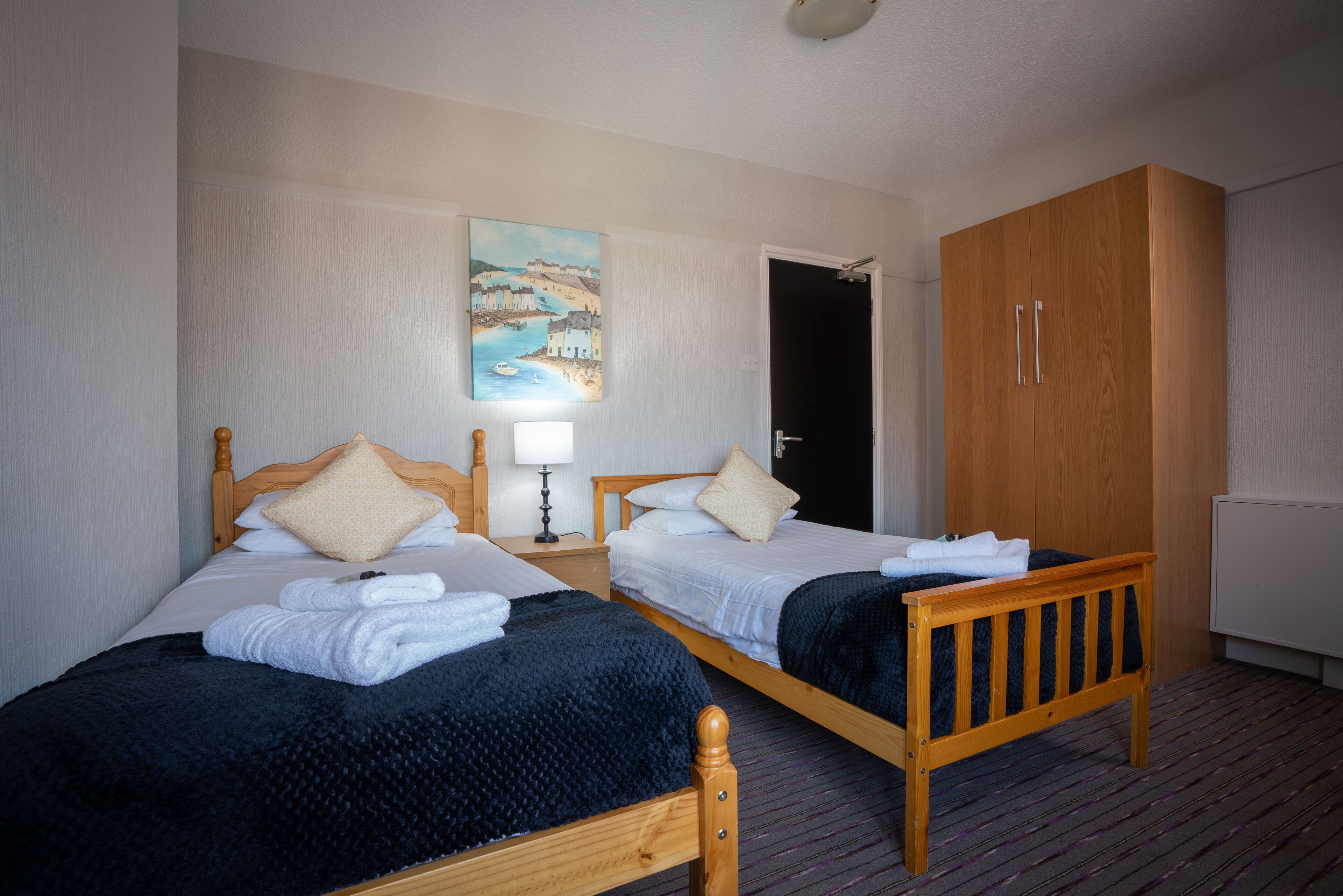 Una habitación de hotel con dos camas de madera, una mesita de noche con lámpara, un armario y un cuadro en la pared.