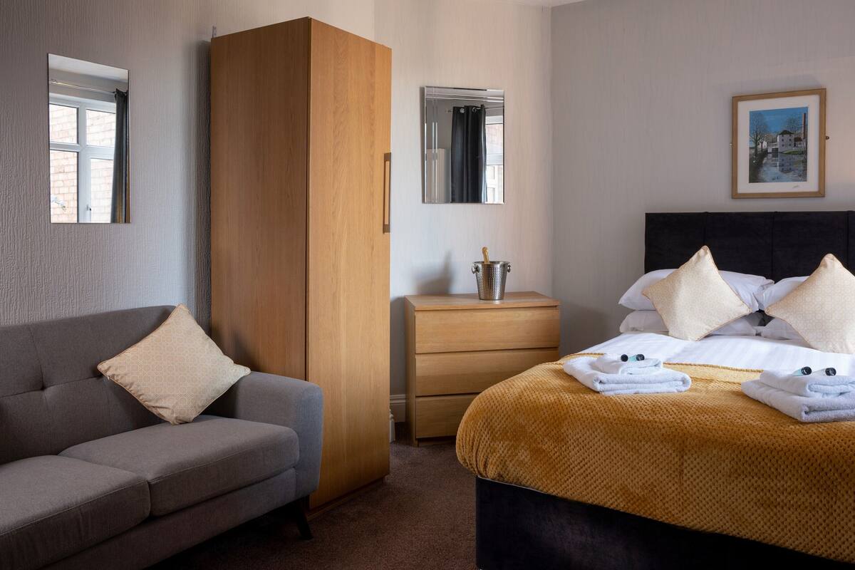 A hotel room with a sofa, a bed with a brown blanket, a nightstand with a lamp, a wooden wardrobe, and a mirror.