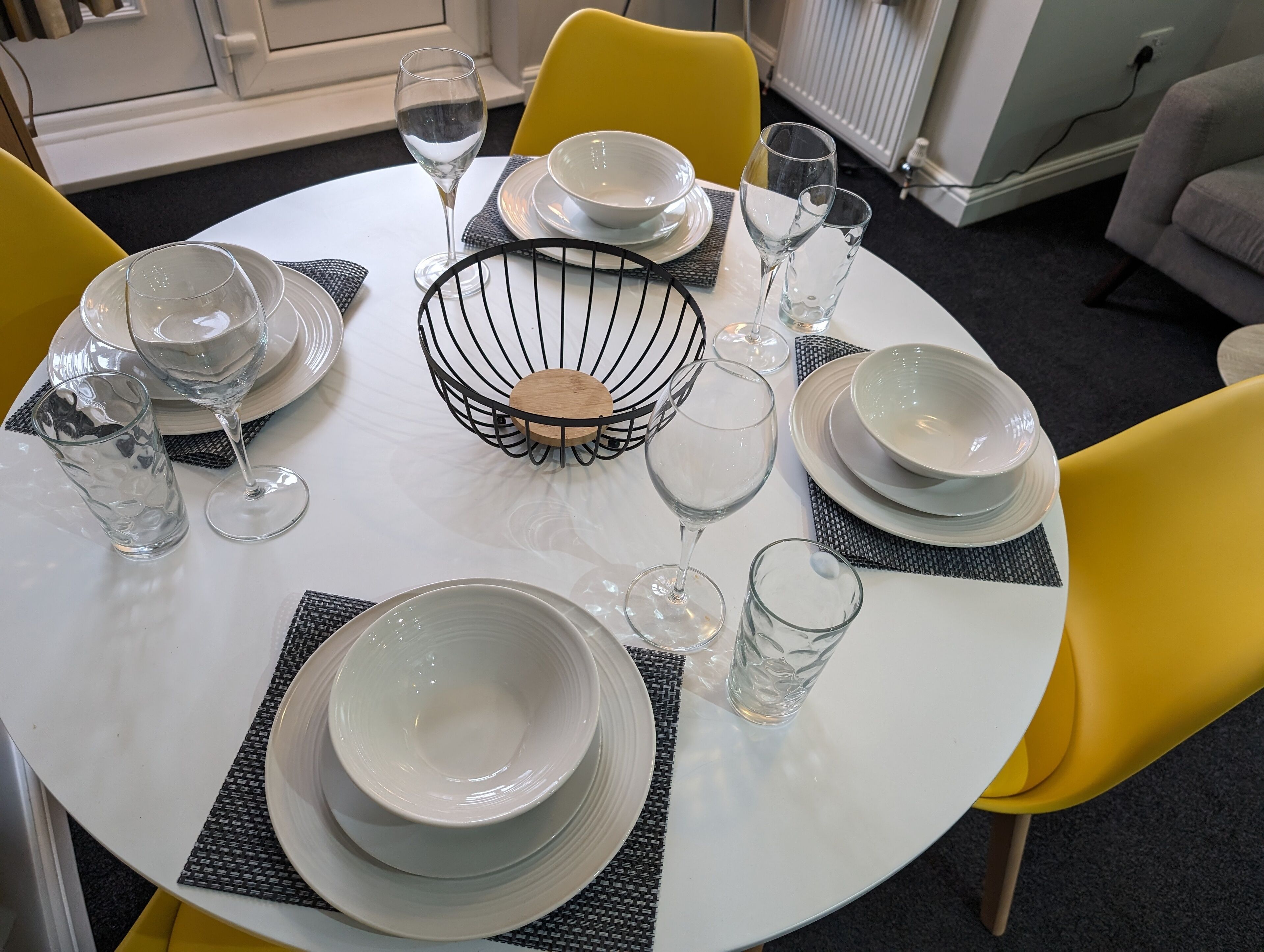 Une table de salle à manger ronde, recouverte d’une nappe blanche, avec des assiettes, des bols et des verres transparents, entourée de chaises jaunes.