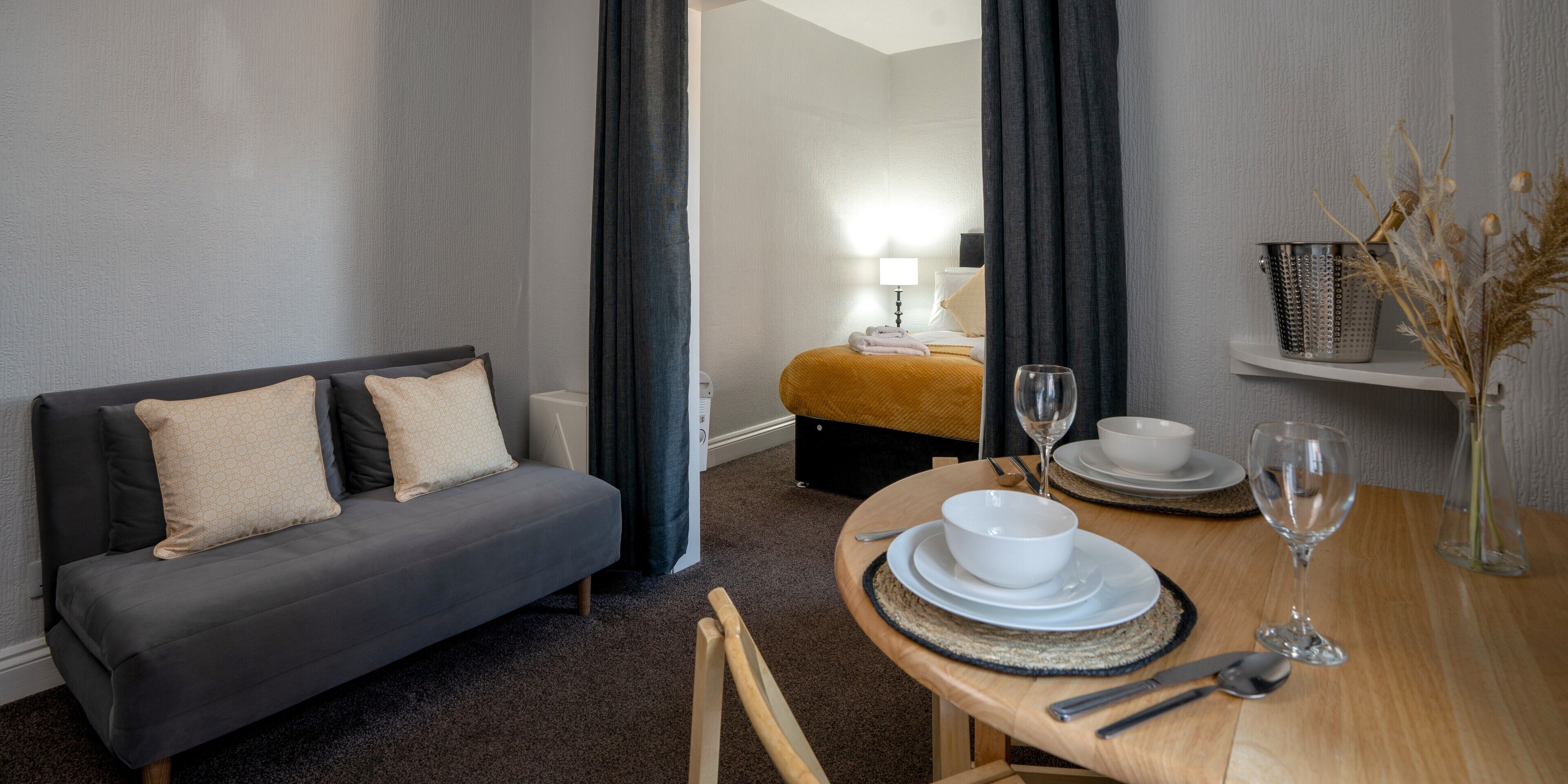 A hotel room with a grey sofa, a wooden dining table set with plates, glasses, and cutlery, and a small shelf with decorative items.