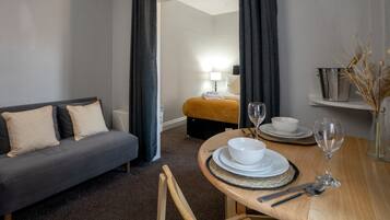 A hotel room with a grey sofa, a wooden dining table set with plates, glasses, and cutlery, and a small shelf with decorative items.