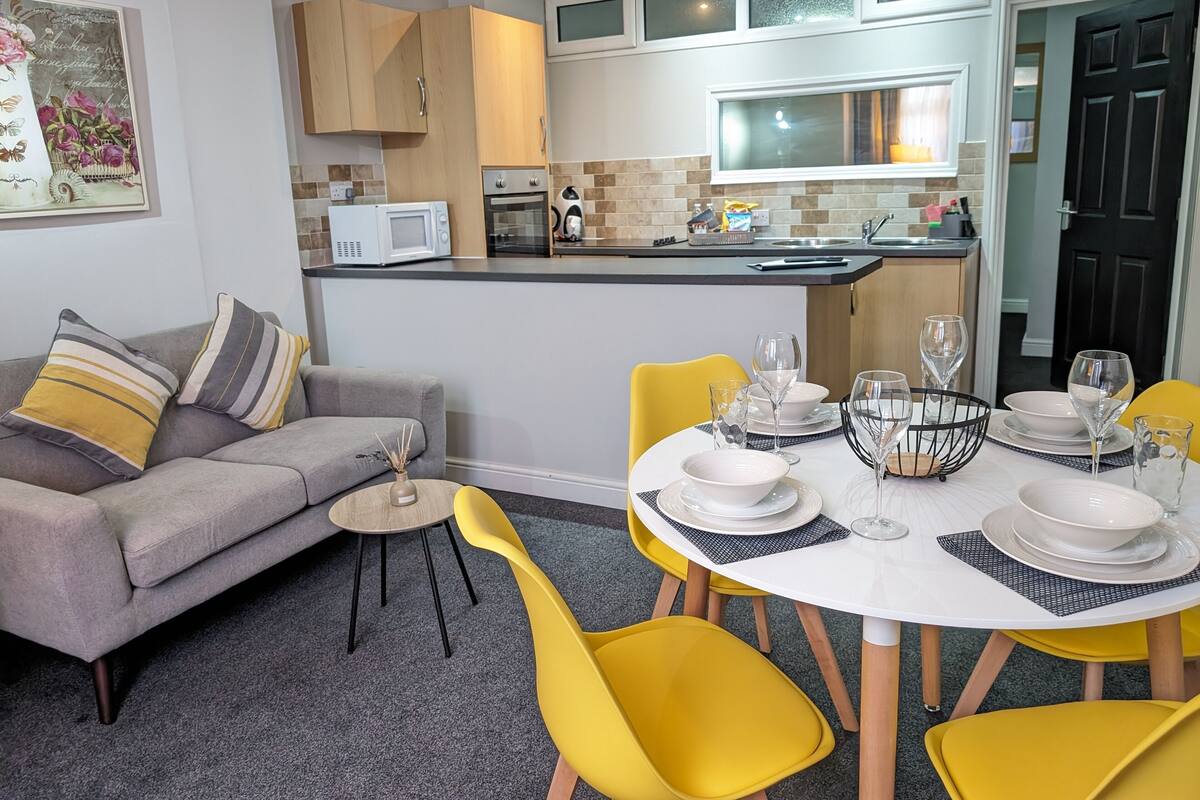 A modern living room with a grey sofa, yellow chairs, and a dining table set for six.