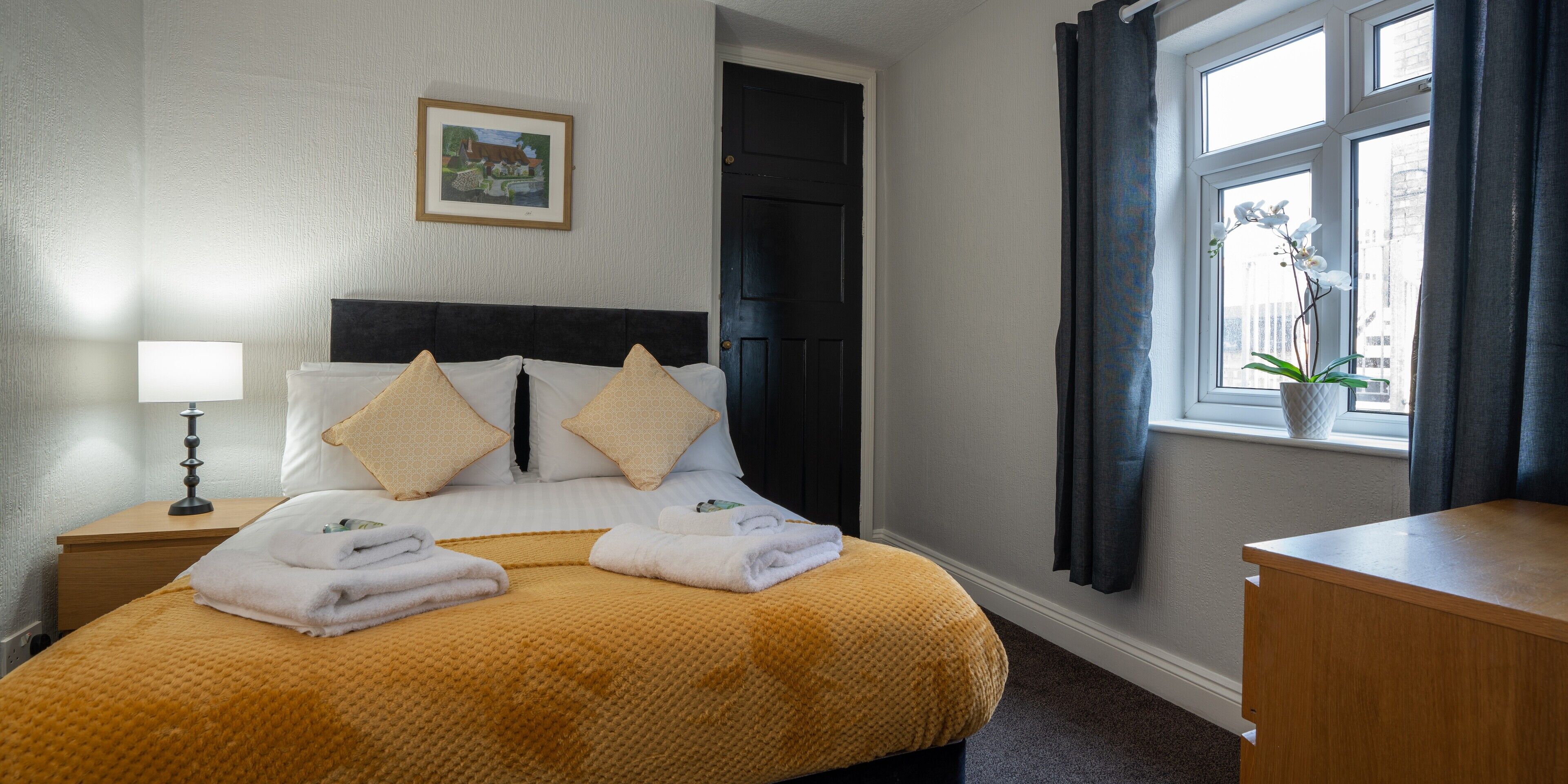 A neatly made bed with a yellow blanket, two pillows, and folded towels. A framed picture hangs on the wall above the bed.