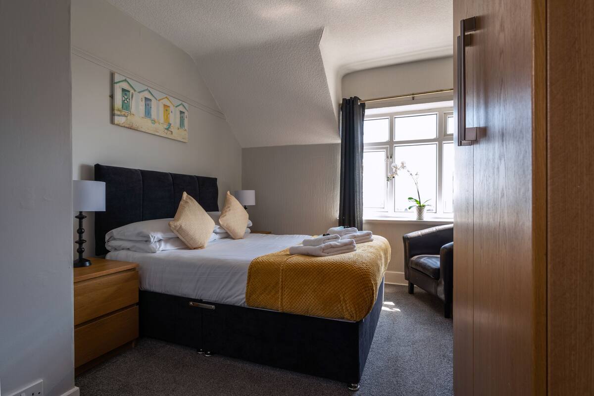 A neatly arranged bedroom with a bed, bedside tables, a lamp, a window with curtains, and a painting on the wall.