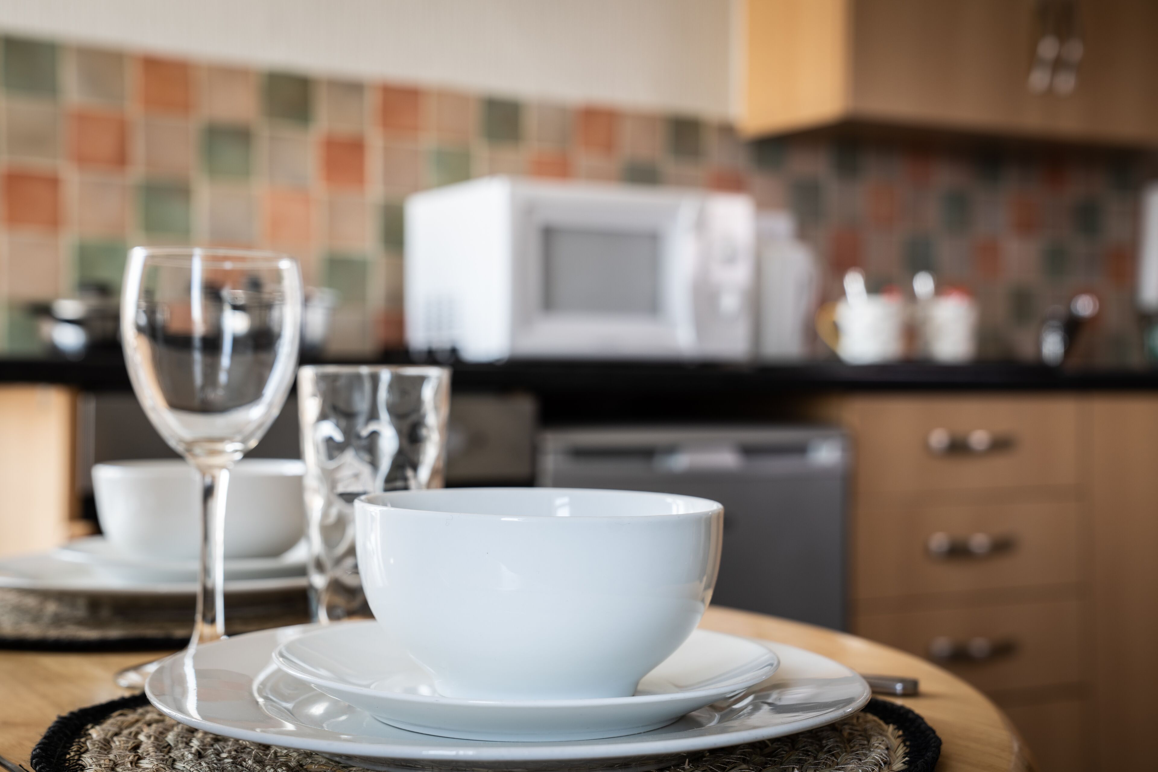 A dining table with a white bowl and plate, a wine glass, and a clear glass. A microwave is visible in the background.