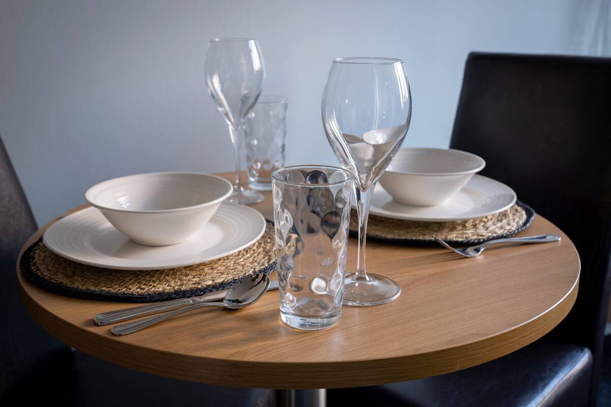 A round dining table with a woven placemat, two white bowls, two clear wine glasses, a clear glass, and cutlery.