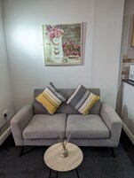 Un sofá gris con cojines a rayas, una mesita redonda con un jarrón y una obra de arte floral en la pared.