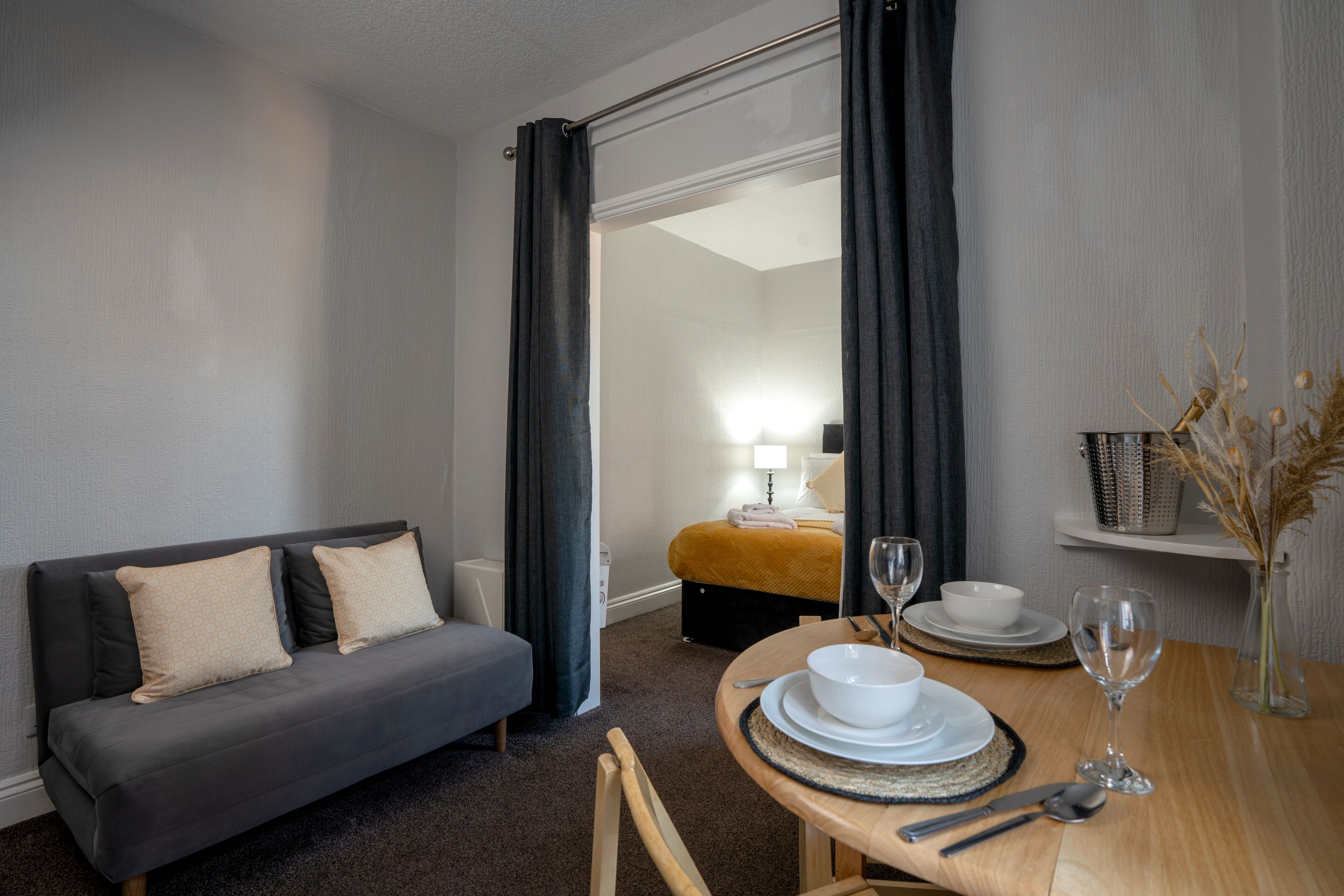 Une chambre d’hôtel avec un canapé gris, une table à manger avec des assiettes, des verres et des couverts, et un petit vase contenant des plantes séchées.