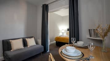 Une chambre d’hôtel avec un canapé gris, une table à manger avec des assiettes, des verres et des couverts, et un petit vase contenant des plantes séchées.