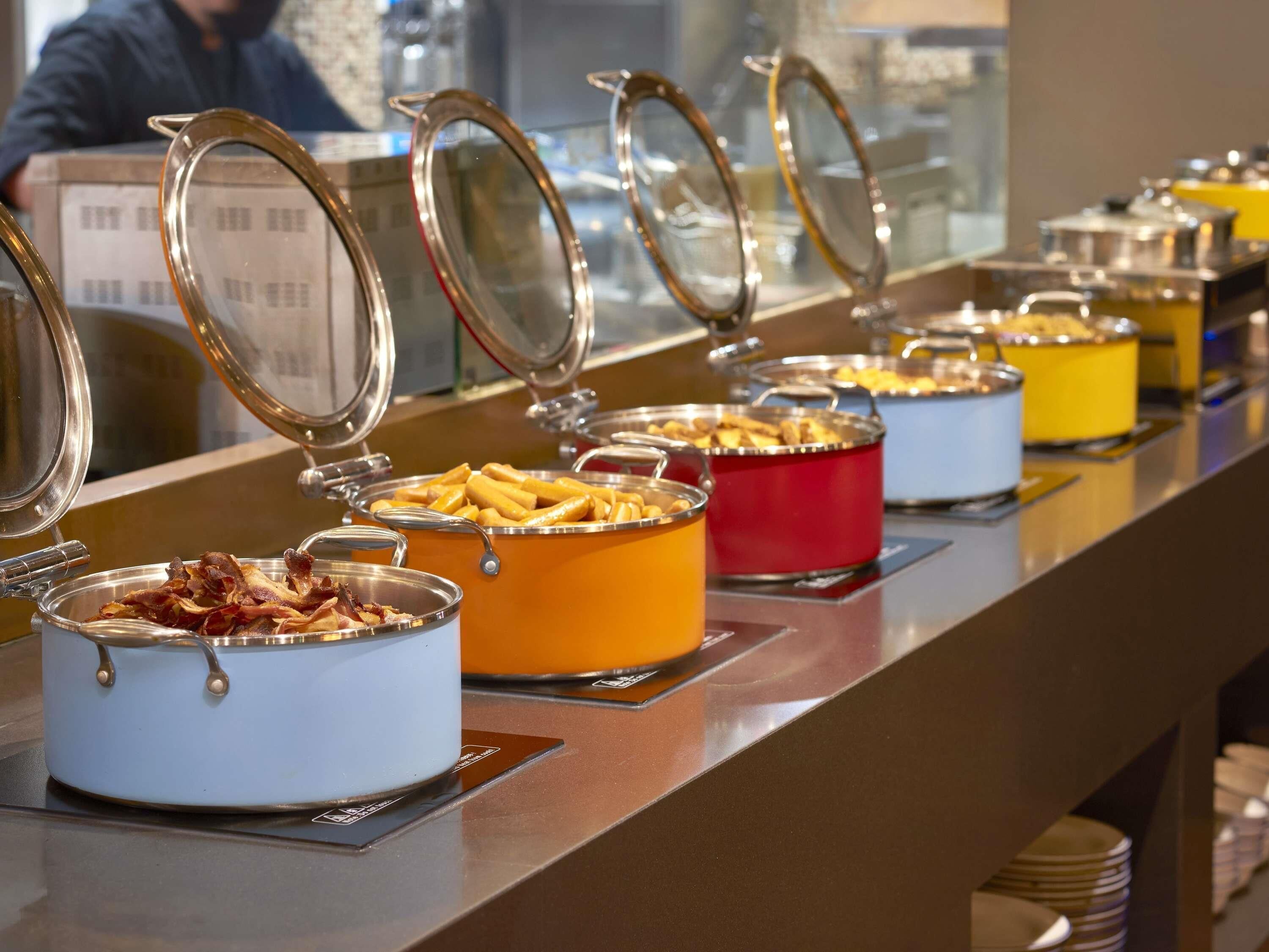 A buffet line with various food items in pots and bowls, a person in the background, and a counter with stacked plates.