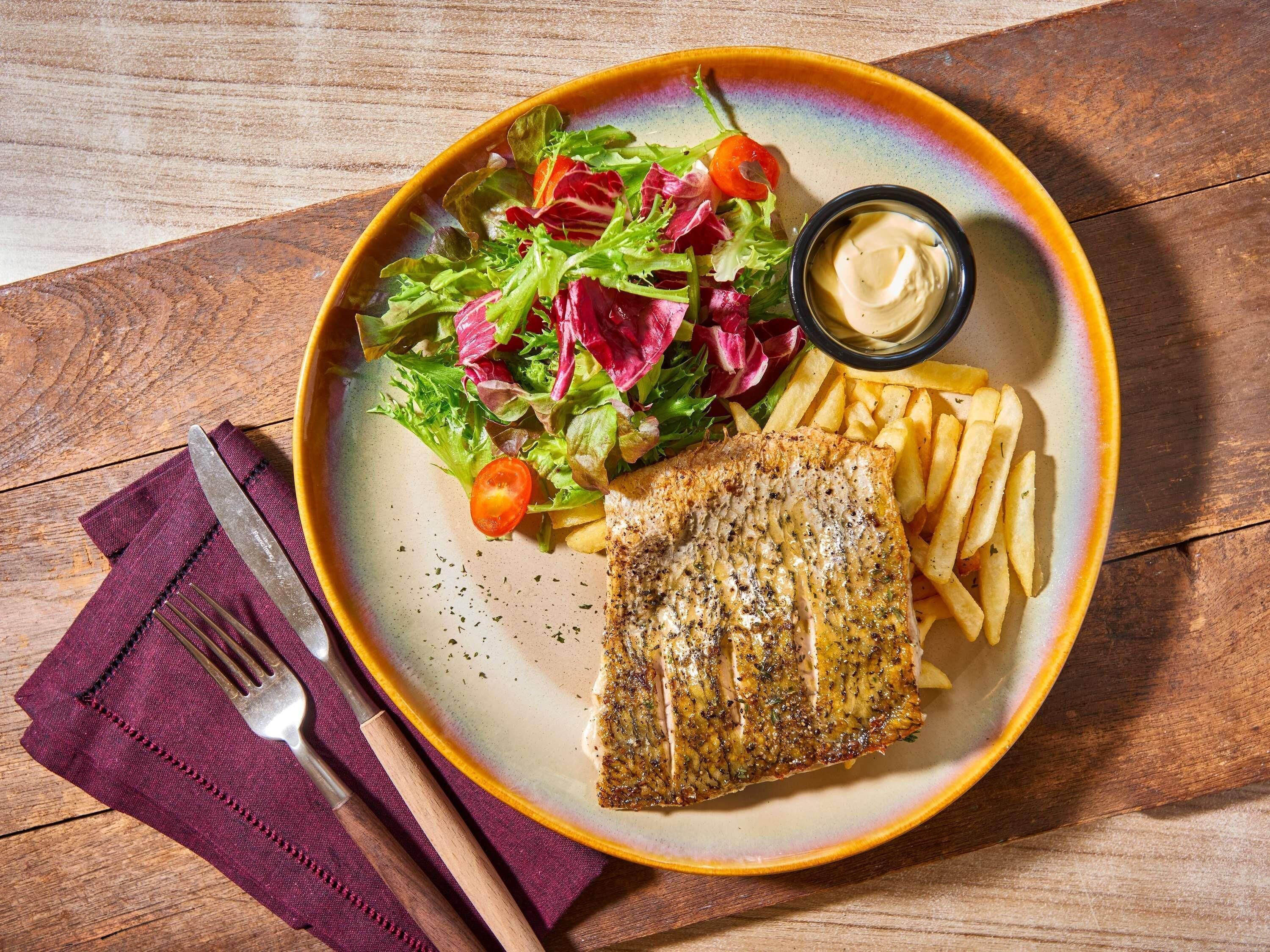 Un plato con milanesa de pescado, papas fritas y una ensalada sobre una mesa de madera.