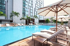 Un espace piscine moderne, avec des chaises longues, des parasols et une vue sur la façade de l’immeuble.