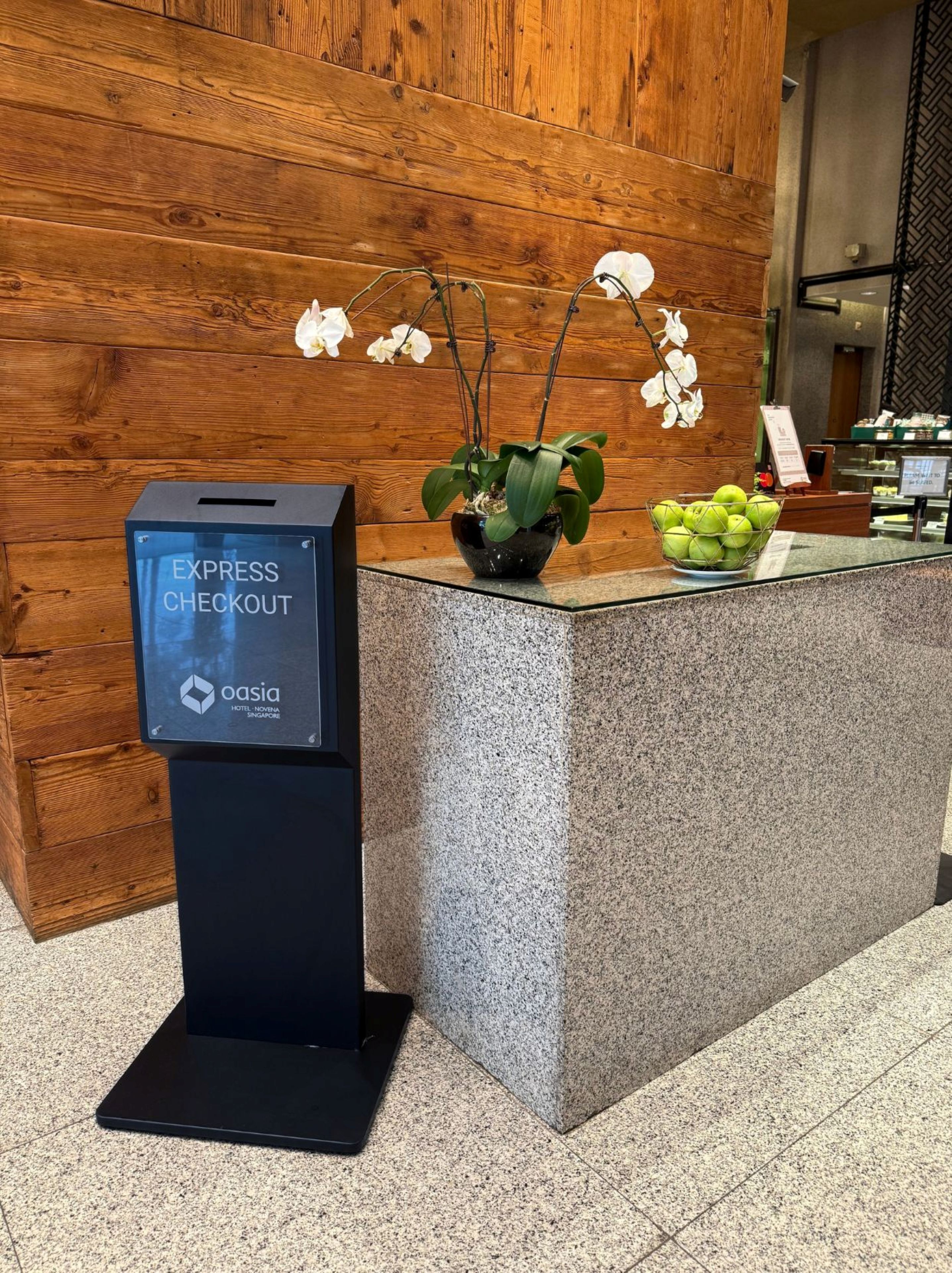 Zona de recepción con pared de madera, una máquina de autopago negro, una planta en maceta y un bol de manzanas verdes.