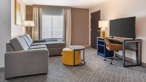 A hotel room with a sofa, a small round table, a desk with a chair, a television, and a window with curtains. - Comfort Inn & Suites Frisco East - McKinney (Frisco)