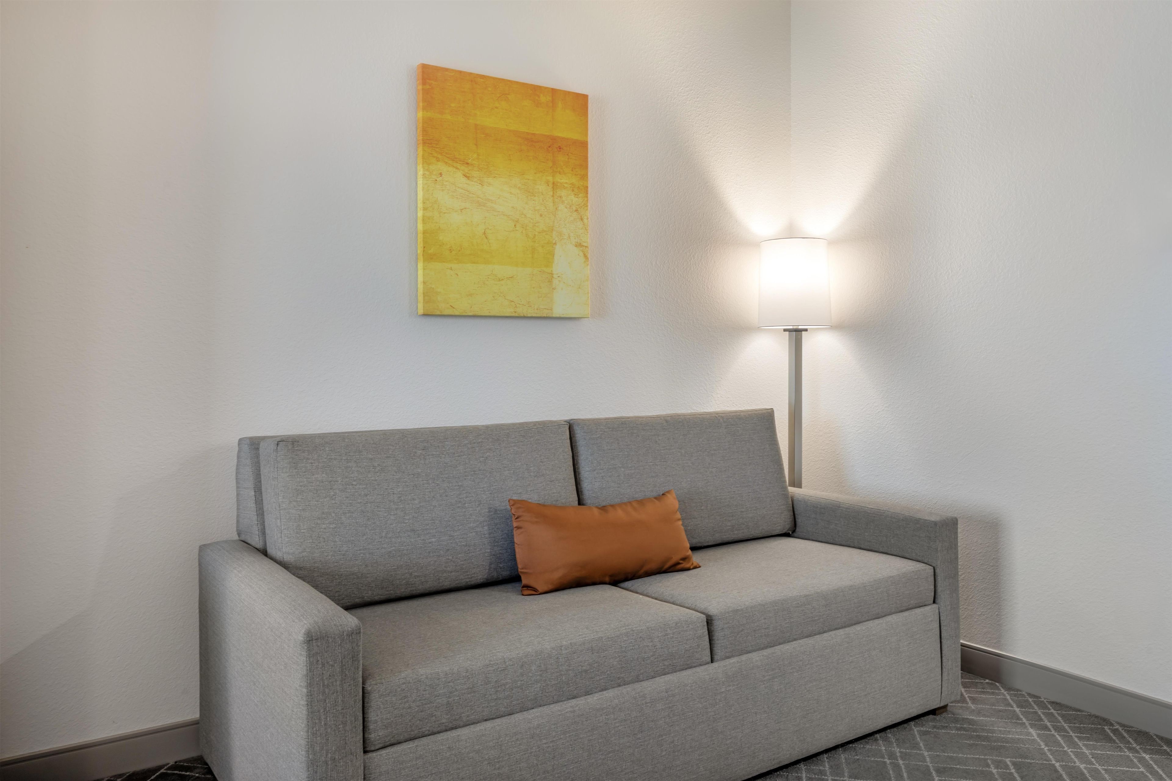 A grey sofa with a brown cushion against a white wall, a floor lamp, and a yellow abstract painting.