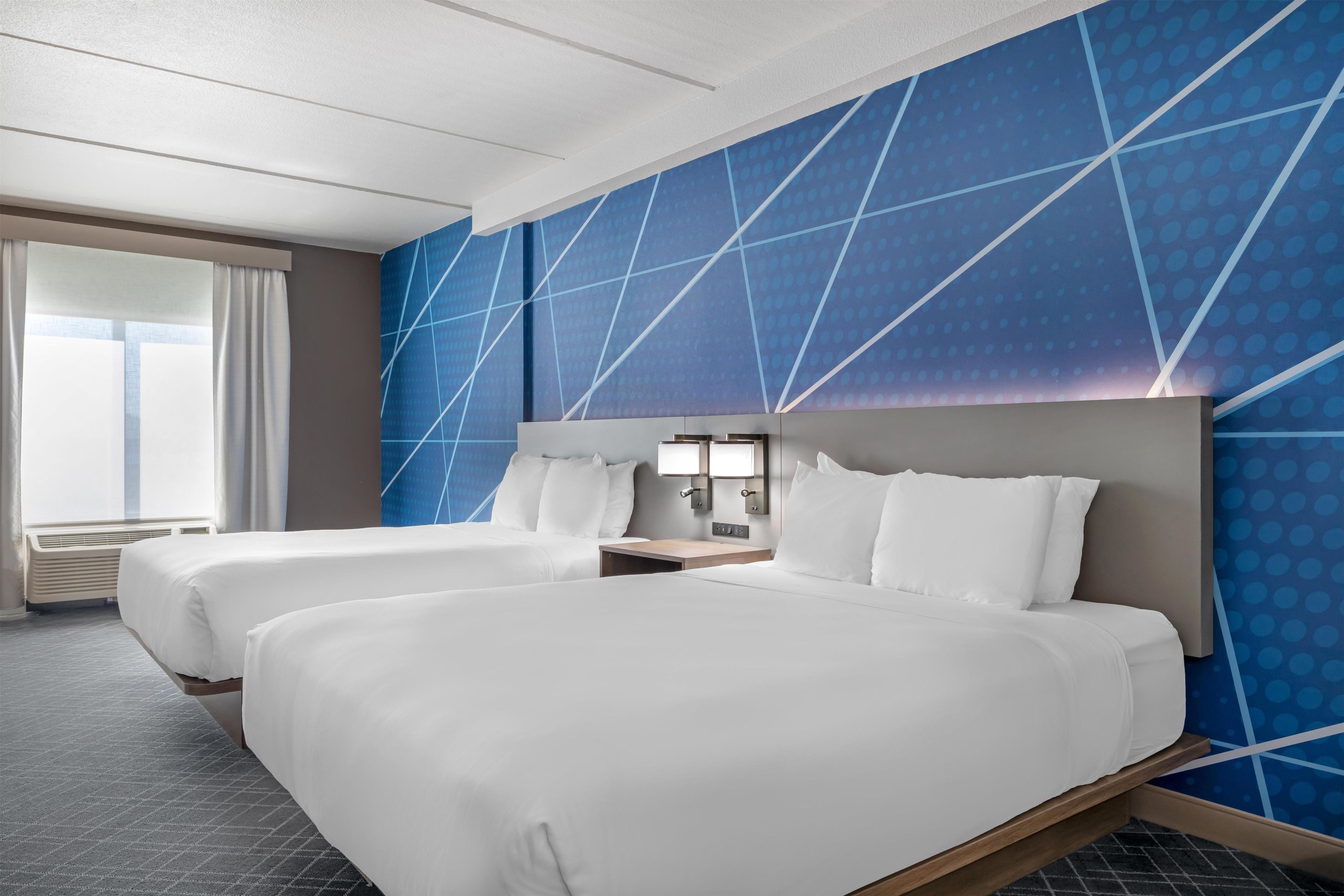 A hotel room with two beds, a large window with curtains, and a blue patterned wall.