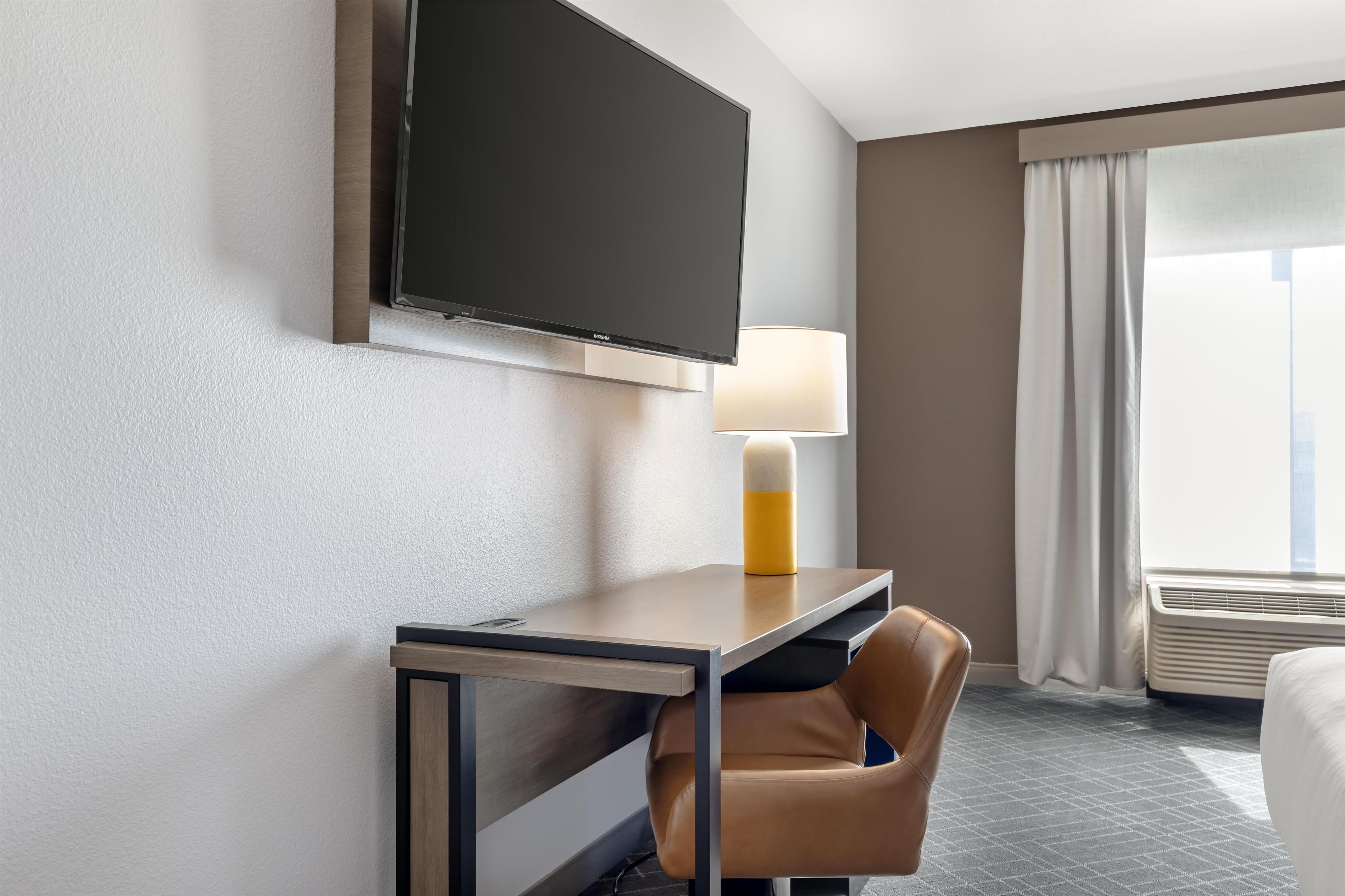 Habitación de hotel con televisor de pantalla plana montado en la pared, un escritorio con lámpara, un sillón de cuero y un ventanal con cortinas.