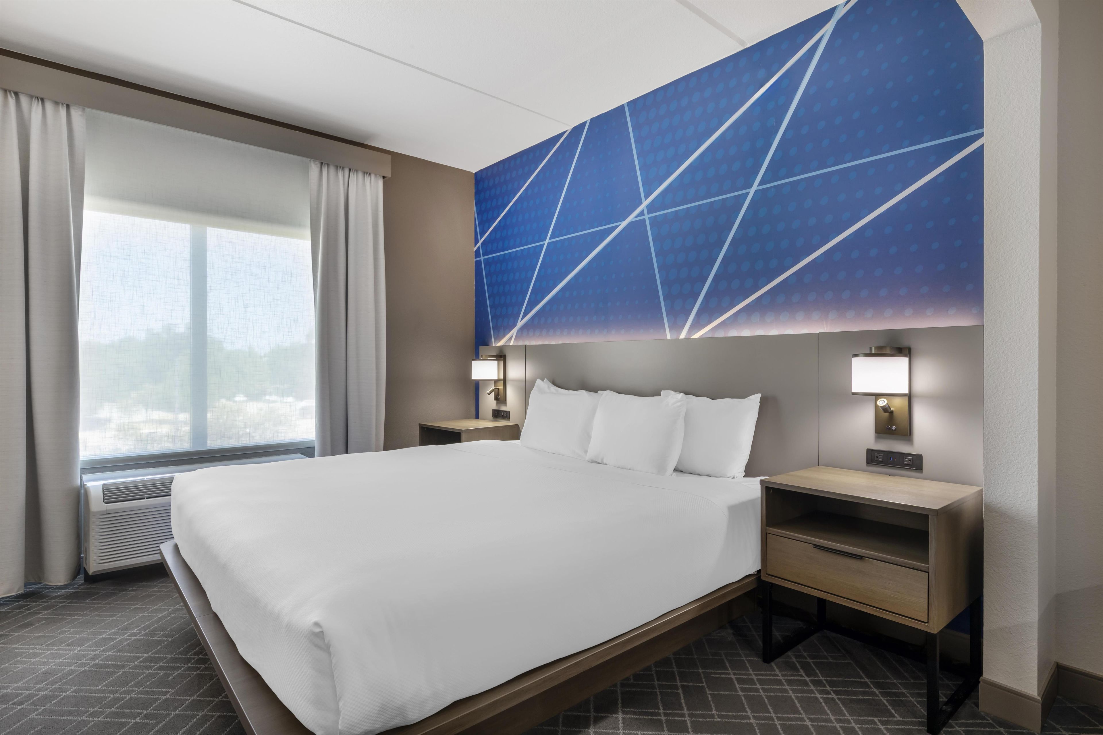 Habitación de hotel con una cama grande, mesitas de noche, una lámpara de pared y un ventanal con cortinas.