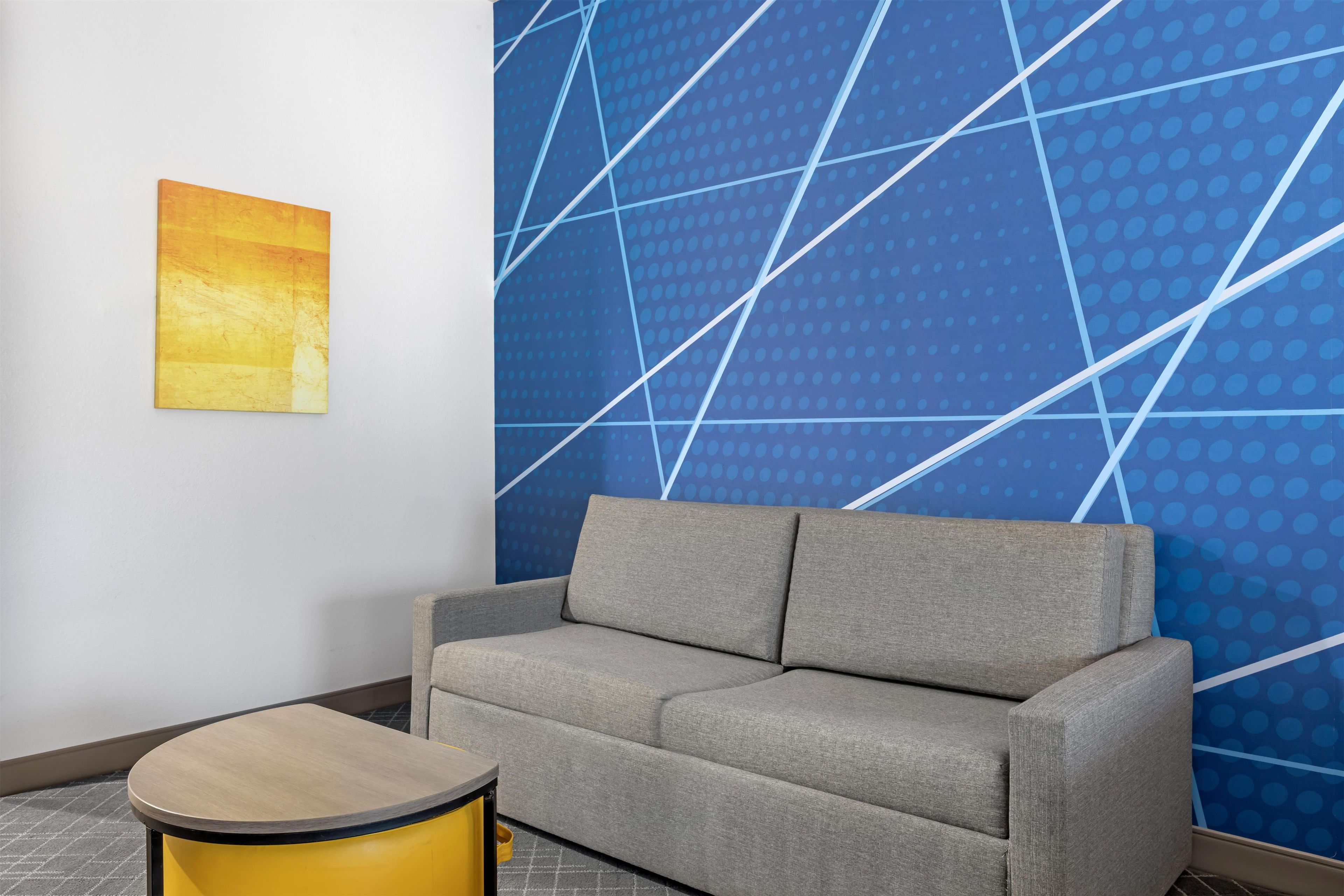 Una habitación moderna con un sofá gris, una mesita auxiliar amarilla y una pared azul con un diseño geométrico.