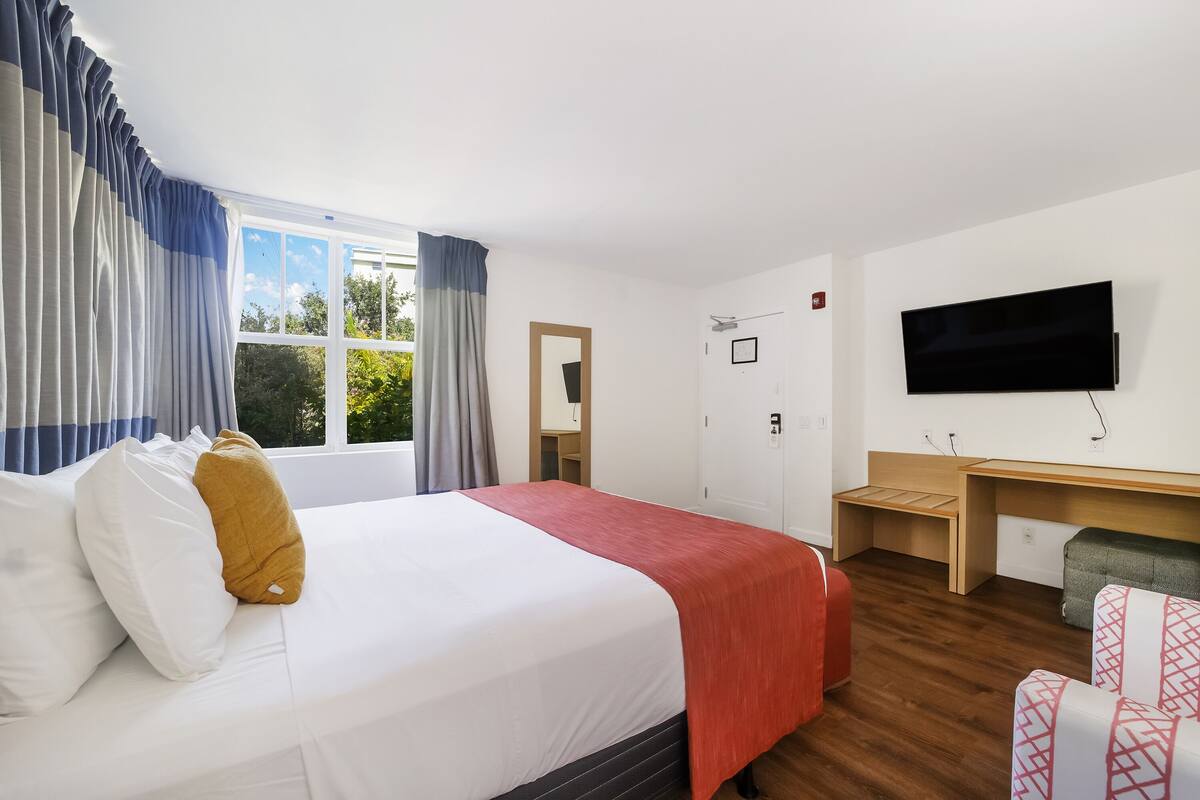 Una habitación de hotel con cama, escritorio, televisor y ventana con cortinas.
