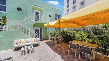 Un patio con un edificio verde, mobiliario exterior y un paraguas amarillo.