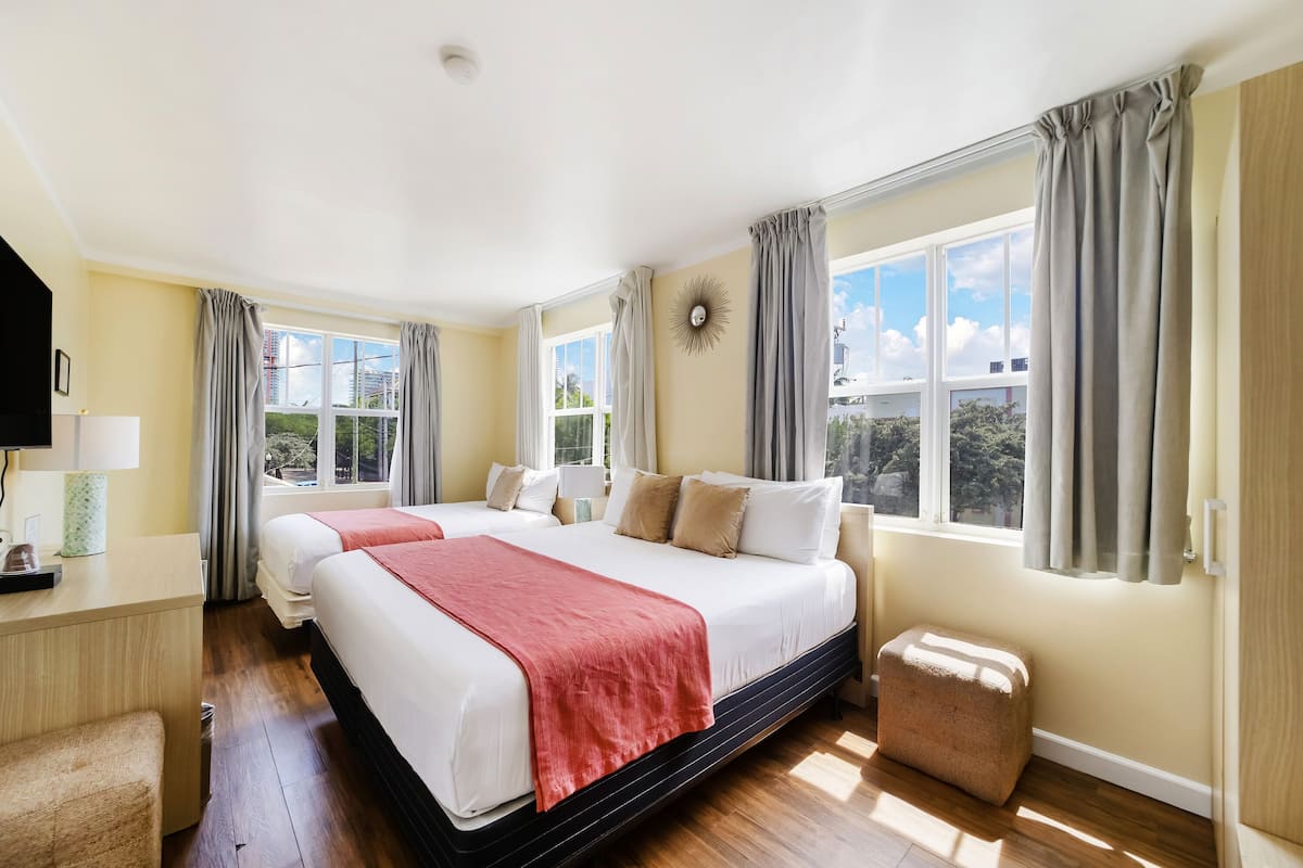 Habitación de hotel con dos camas, un escritorio, una silla, un pequeño reposapiés y ventanas grandes con cortinas.
