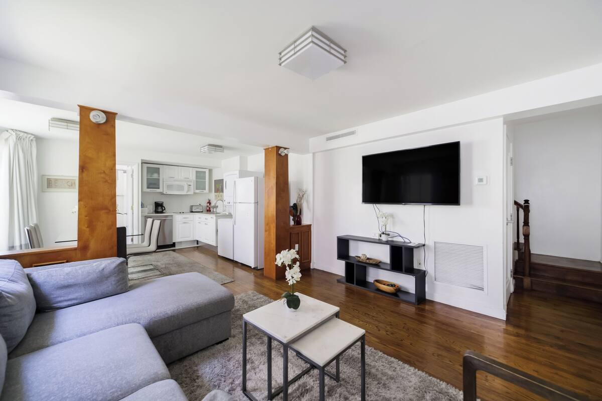 Una sala moderna con un sofá seccional gris, una pequeña mesa blanca y un televisor de pantalla plana montado en la pared. Se ve una cocina al fondo, con gabinetes blancos y un microondas.