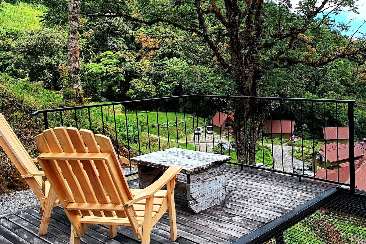 Una terraza de madera con una mesa y sillas, con vistas a un paisaje verde exuberante con casas y una montaña al fondo.