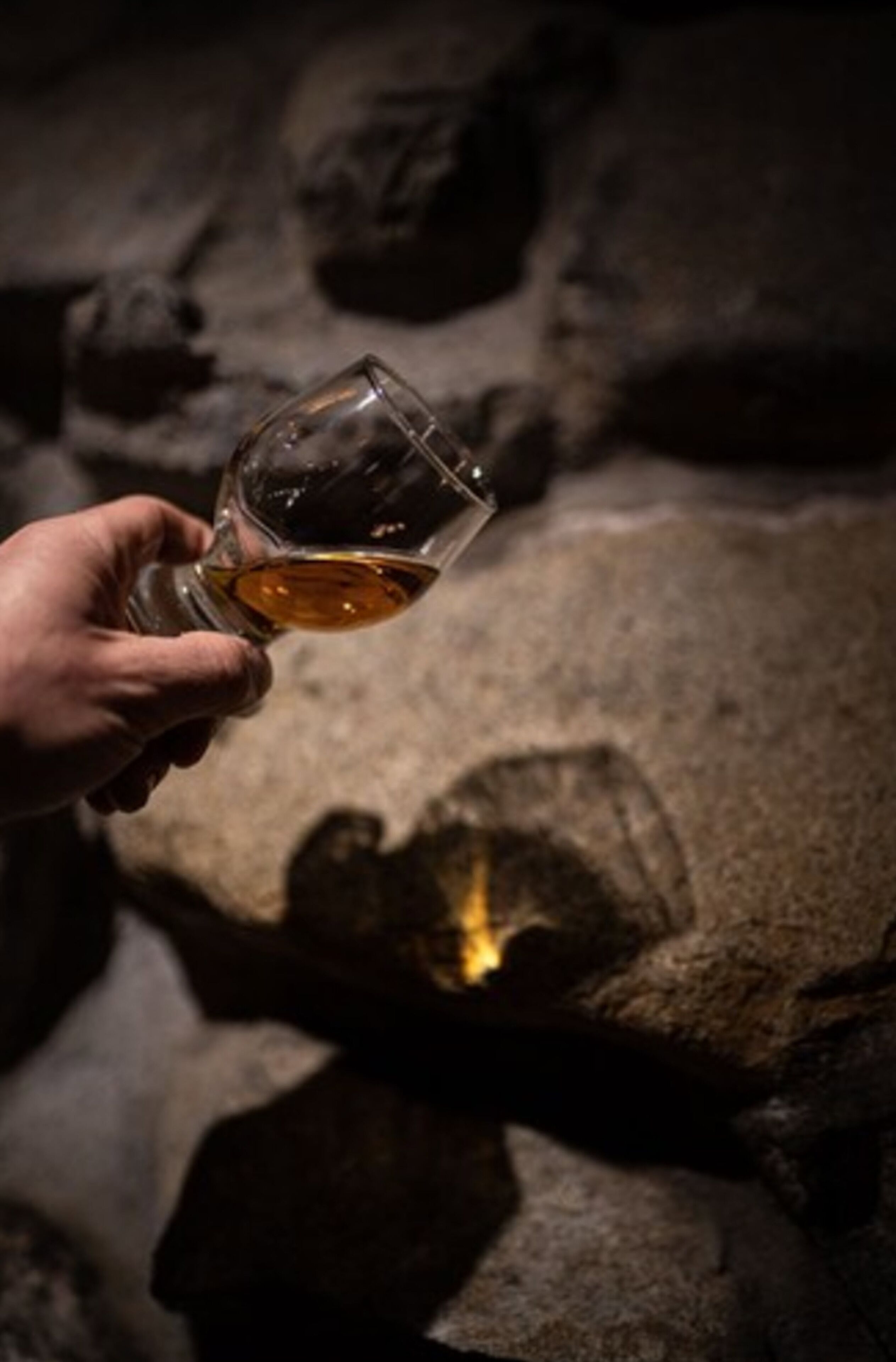 A hand holding a glass of amber liquid, possibly whiskey, with a shadow of a person on a stone surface.